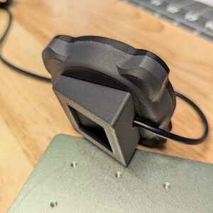 Op de afbeelding: Een zwarte 3D-geprinte camerabevestiging met een vierkante basis en een ronde bovenkant. De bevestiging is verbonden met een zwarte kabel.