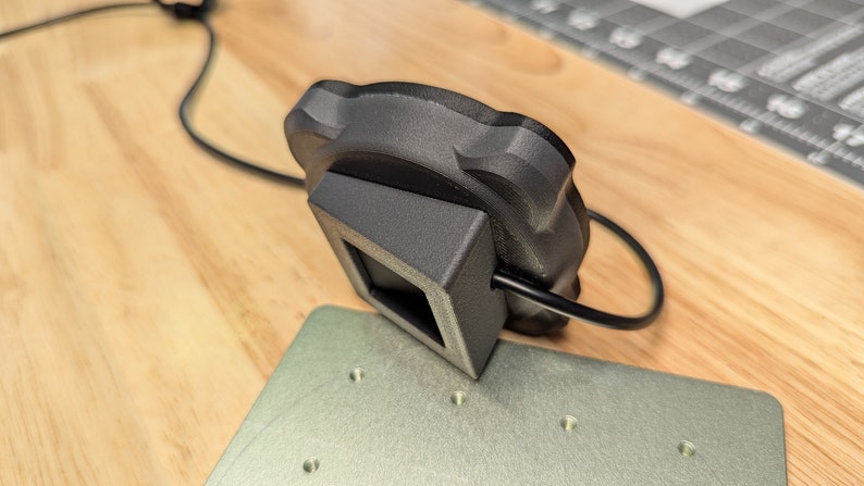 Op de afbeelding: Een zwarte 3D-geprinte camerabevestiging met een vierkante basis en een ronde bovenkant. De bevestiging is verbonden met een zwarte kabel.