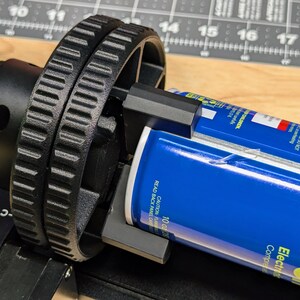 K&ouml;nnte beinhalten: Eine schwarze und silberne Dosenrolle mit einer blauen Dose, die eingesetzt ist. Die Rolle hat einen schwarzen Riemen und ein schwarzes Zahnrad. Die Dose ist mit "Super Oil Electric Contact Cleaner" beschriftet.