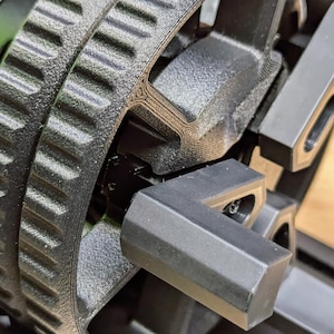 K&ouml;nnte beinhalten: Nahaufnahme eines schwarzen, 3D-gedruckten Zahnrads mit einem Zahnriemen und einer schwarzen Kunststoffhalterung. Das Zahnrad ist an einer schwarzen Metallwelle befestigt.