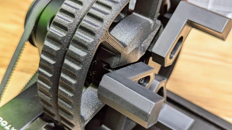 K&ouml;nnte beinhalten: Nahaufnahme eines schwarzen, 3D-gedruckten Zahnrads mit einem Zahnriemen und einer schwarzen Kunststoffhalterung. Das Zahnrad ist an einer schwarzen Metallwelle befestigt.