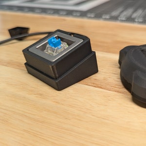 Op de afbeelding: Twee 3D-geprinte zwarte kunststof schakelaarkasten. Een behuizing heeft een blauwe en zilveren mechanische toetsenbordschakelaar ge&iuml;nstalleerd.