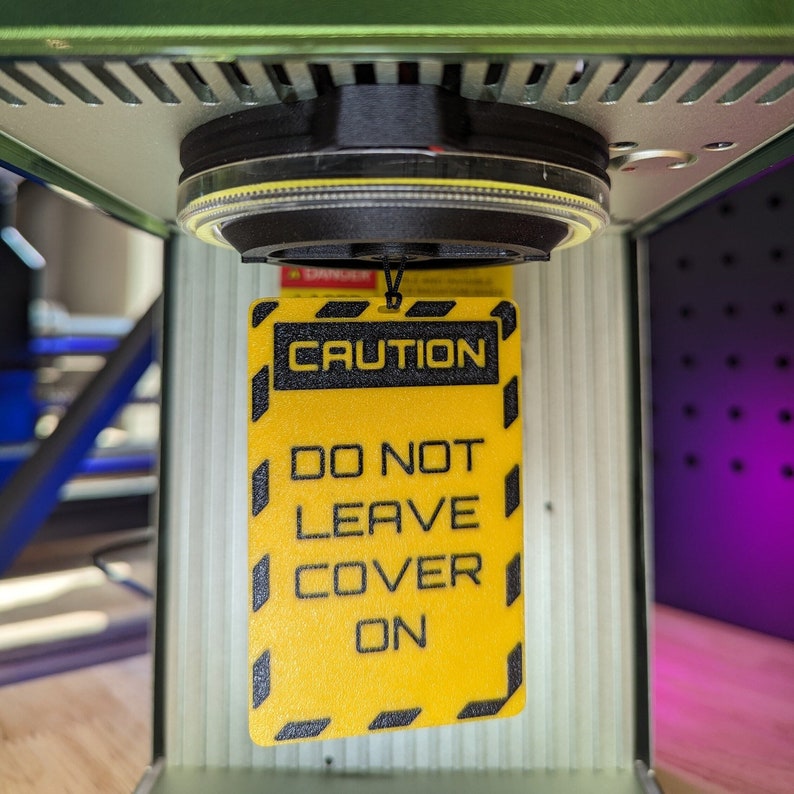 Op de afbeelding: Een geel waarschuwingsbord met zwarte tekst die "CAUTION DO NOT LEAVE COVER ON" luidt. Het bord hangt aan een zwart en zilver apparaat.