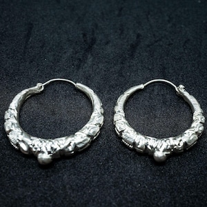 Pode incluir: Um par de brincos de argola prateados com um design ornamentado e texturizado. Os brincos têm formato circular com um padrão detalhado ao longo da borda externa e um pequeno adorno arredondado na parte inferior. O fundo é escuro.