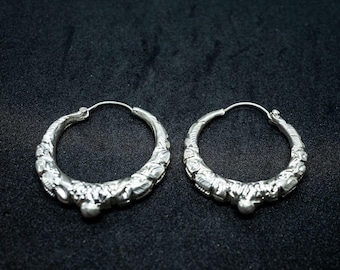 May include: A pair of silver hoop earrings with an ornate, textured design. The earrings have a circular shape with a detailed pattern along the outer edge and a small, rounded embellishment at the bottom. The background is a dark, solid color.