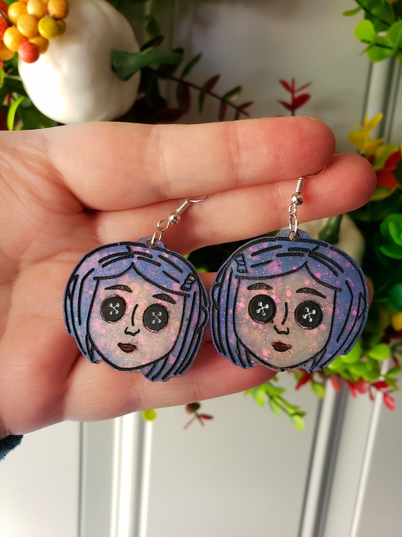 Puede incluir: Un par de pendientes de purpurina azul y morado con un personaje de dibujos animados con cabello azul y ojos de bot&oacute;n. La cara del personaje es rosa y blanca.