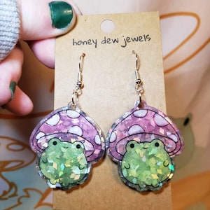 May include: A pair of iridescent green frog earrings with pink mushroom caps. The earrings are made of resin and have a glittery finish. The earrings are on a brown card with the text "honey dew jewels".
