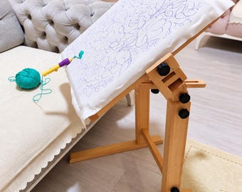 May include: A wooden floor stand holding a white fabric frame with a blue floral outline pattern. A punch needle tool with a teal yarn ball is positioned on the fabric. The stand features adjustable joints with black knobs for height and angle.