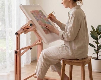 May include: A wooden floor-standing embroidery frame with an adjustable tilt mechanism, holding a rectangular fabric canvas. The frame features multiple black adjustment knobs and a sturdy base, designed to support needlework projects.