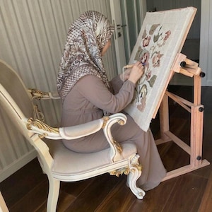 May include: A punch needle embroidery project featuring a floral design on a rectangular fabric canvas mounted on a wooden floor stand. The embroidery displays various shades of pink, green, and cream yarn, worked with a handheld punch needle tool.