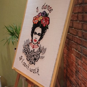 Puede incluir: Obra de arte bordada sobre lienzo que representa a una mujer con un tocado floral y el texto "I dance if I can't walk". La obra presenta flores rojas, naranjas, amarillas y moradas, con detalles en negro y gris. El lienzo está enmarcado en madera clara y expuesto en un caballete de madera.