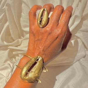 May include: Gold-toned jewelry set featuring a ring and bracelet, both with a seashell design. The ring is worn on a finger, and the bracelet is on a wrist. The jewelry has a polished finish and a natural, organic aesthetic.
