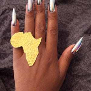 May include: A hand with long, metallic silver stiletto nails and a textured gold ring in the shape of the African continent. The ring is worn on the ring finger. The nails have a reflective, chrome-like finish.