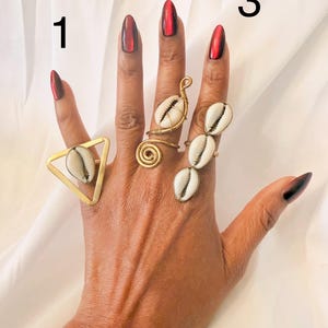 May include: A hand displaying three unique rings. The rings feature cowrie shells and gold-colored metal. One ring has a triangular design, another is a spiral, and the third has three shells. The nails are painted with a dark red ombre.