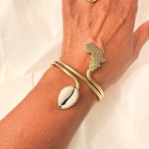 May include: Gold-toned bracelet featuring a cowrie shell and an African continent design. The bracelet wraps around the wrist with a double band. A spiral ring is also visible.