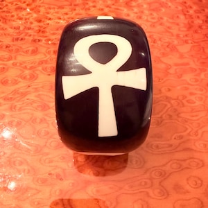 May include: A dark, rounded object with a white ankh symbol. The ankh is a cross with a loop at the top, a symbol of life and immortality. The object is set against a reddish-orange, patterned surface.