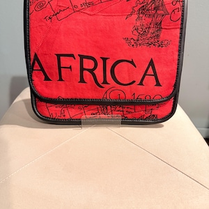 May include: A red messenger bag with black trim featuring a black printed map design and the word AFRICA in large capital letters. The flap includes a black line drawing of a sailing ship and the text 1500, with black piping along the edges.
