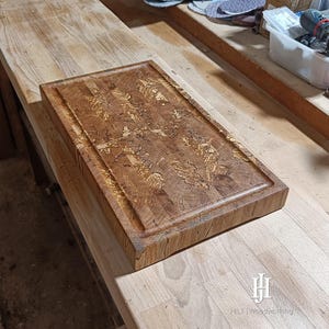 Könnte beinhalten: Ein rechteckiges Holzschneidebrett mit Saftrille. Das Brett besteht aus hellem und dunklem braunem Holz mit einem einzigartigen Muster. Die Kanten sind leicht erhöht und das Gesamtdesign ist rustikal und handgefertigt. Das Logo "HILT Woodworking" ist unten rechts sichtbar.