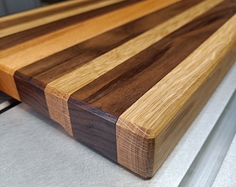 May include: A rectangular cutting board constructed from alternating strips of dark brown walnut wood and lighter tan oak wood. The board features a smooth, polished surface with visible wood grain patterns and beveled edges on the corners.