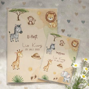 Könnte beinhalten: Zwei beige Baby-Aufnahmebücher mit Safari-Tierillustrationen. Das größere Buch trägt die Aufschrift "U-Heft" und das kleinere Buch "Impfpass". Beide Bücher tragen den Namen "Lio Kurz" und das Datum "25 JULI 2022".