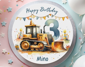 May include: A round, silver cake topper with a white background. The topper features a yellow bulldozer with a blue number 3, surrounded by colorful flags and candles. The text "Happy Birthday" and "Mino" are printed on the topper.