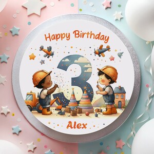 May include: A white round cake with a silver border. The cake is decorated with a number 3, two children wearing hard hats, a toy construction truck, and a toy block tower. The cake says "Happy Birthday Alex" in orange and white text.