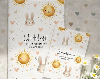May include: Two white baby record books with a repeating pattern of smiling suns, bunnies, hearts, and stars. The larger book reads "U-Heft" and the smaller book reads "Impfpass". Both books have the name "LUNA SCHMIDT" and the date "25 MAI 2022".