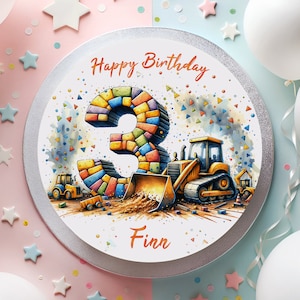 Op de afbeelding: Een ronde witte taart met een zilveren rand. De taart is versierd met een kleurrijk nummer drie gemaakt van blokken, een geel bouwvoertuig en confetti. De tekst "Happy Birthday" en "Finn" staat op de taart.