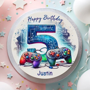 Könnte beinhalten: Ein runder Kuchen mit silbernem Rand und weißem Hintergrund. Der Kuchen ist mit einer blauen und violetten Zahl 5, zwei bunten Videospielcontrollern und den Worten "Happy Birthday" und "Justin" verziert.