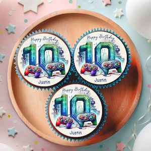 Könnte beinhalten: Drei runde Cupcakes mit blauem Frosting und essbaren Bildern. Jeder Cupcake hat ein farbenfrohes Bild eines Videospiel-Controllers und die Zahl 10. Der Text "Happy Birthday" und "Justin" ist auf jedem Cupcake gedruckt.