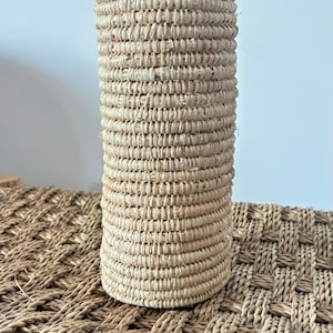 Op de afbeelding: Een natuurlijke geweven mand met een cilindrische vorm en een lichtbruine kleur. De mand is gemaakt van strak geweven strengen van natuurlijk materiaal, waardoor een textuur ontstaat.