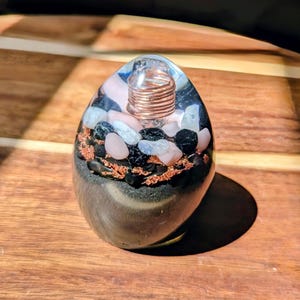 Puede incluir: Un generador de energía orgónica en forma de huevo. La resina transparente encierra una bobina de cobre, piedras negras, blancas y rosas, y copos de cobre. La base es de color gris oscuro. El objeto está sobre una superficie de madera.