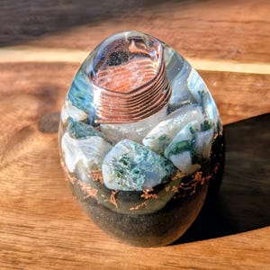 Puede incluir: Un pisapapeles ovalado de resina con una bobina de cobre y piedras de ágata musgosa. La resina transparente encierra la bobina de cobre y las piedras, con una base oscura. Las piedras tienen patrones verdes y blancos. El pisapapeles está sobre una superficie de madera.