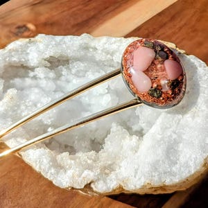 Puede incluir: Una horquilla decorativa con un tallo dorado y una parte superior ovalada. La parte superior presenta una mezcla de piedras rosas y marrones y una base color cobre. El accesorio para el cabello está sobre una superficie blanca similar a un cristal.