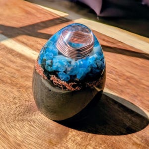 Puede incluir: Una pieza de arte de resina en forma de huevo con una base oscura, inclusiones de cristales azules y detalles de alambre de cobre. Una parte superior de resina transparente encierra una bobina de cobre. La pieza está sobre una superficie de madera.
