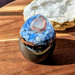 Puede incluir: Una pieza de arte de resina de forma ovalada con una bobina de alambre de cobre y un cristal transparente en la parte superior. La pieza presenta piedras azules y blancas encapsuladas en resina transparente, con una base marrón oscuro. Una geoda está en el fondo.