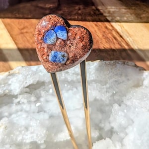 Puede incluir: Una horquilla para el pelo en forma de corazón con una base color cobre e incrustaciones de piedras azules. La horquilla tiene dos púas doradas. El corazón mide aproximadamente 3,8 cm de ancho.