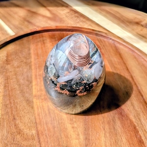 Puede incluir: Una pieza de arte de resina de forma ovalada con una bobina de alambre de cobre en la parte superior. El interior presenta piedras claras y negras, con una base de color cobre. La pieza está colocada sobre una bandeja de madera.