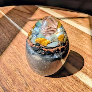 Puede incluir: Un generador de energía orgónica en forma de huevo. La resina transparente encierra una variedad de cristales coloridos, incluyendo ojo de tigre, turmalina negra y cuarzo, junto con alambre de cobre. La base es de color oscuro y metálico.