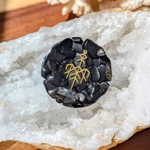 May include: A circular resin coaster or decorative piece filled with small, polished black stones. Embedded in the center is a gold-colored metal charm featuring a geometric, branching symbol resembling a stylized tree or a runic stave design.