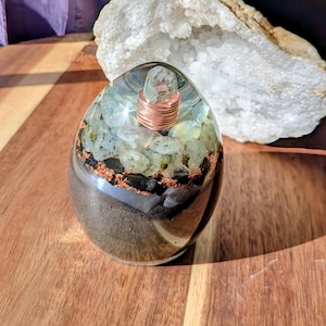Puede incluir: Una pieza de arte de resina transparente en forma de huevo con una bobina de cobre y un cristal en la parte superior. El interior presenta piedras verdes y negras y arena. La pieza está sobre una superficie de madera.
