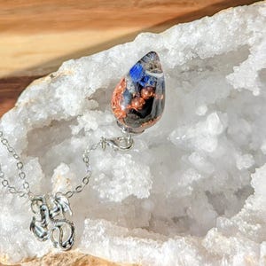 May include: A silver necklace with a pendant featuring a teardrop-shaped resin design with blue, copper, and black elements. The necklace also has a charm with a dragon design. The pendant is approximately 1.5 inches long.