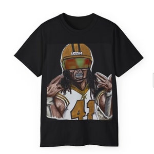 Peut inclure: T-shirt noir avec un imprimé graphique d'un joueur de football portant un casque doré avec le mot "Saints" dessus. Le joueur porte un maillot blanc avec le numéro "41" dessus.