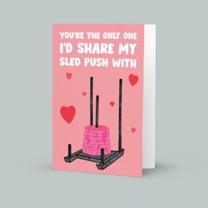 Pu&ograve; includere: Un biglietto di San Valentino rosa con il testo "You're the only one I'd share my sled push with." Il biglietto presenta un'immagine di un trenino con dei pesi sopra.