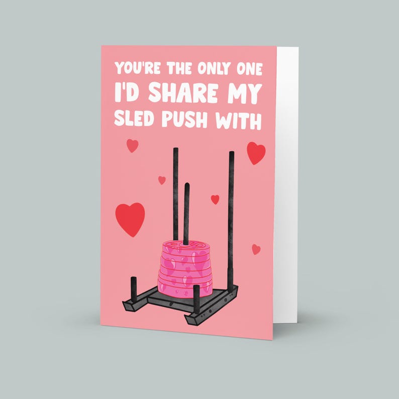 Pu&ograve; includere: Un biglietto di San Valentino rosa con il testo "You're the only one I'd share my sled push with." Il biglietto presenta un'immagine di un trenino con dei pesi sopra.