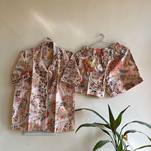 May include: A two-piece pajama set consisting of a short-sleeved button-down shirt with a notched collar and matching elastic-waist shorts. Both pieces feature a light pink base color with a repeating floral pattern in shades of orange, yellow, and green.