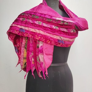May include: A bright pink scarf displayed on a mannequin. The scarf has a layered design with patterned fabric strips, creating a textured look. The edges are finished with fringing. A striking fashion accessory.
