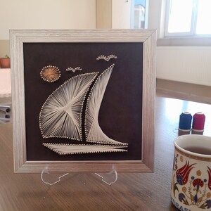 May include: A framed string art piece depicting a sailboat with a sun and birds in the background. The frame is silver with a black mat. The artwork is on a dark brown background.