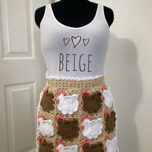 May include: A white tank top with the word "BEIGE" and three hearts printed in brown. Below, a beige crochet skirt with brown and white chicken designs and red accents is displayed on a mannequin.
