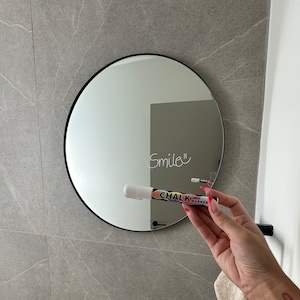 May include: A round, black framed mirror with a white inscription that reads "Smile" on the reflective surface. A white chalk marker with black and colourful lettering is held in front of the mirror.